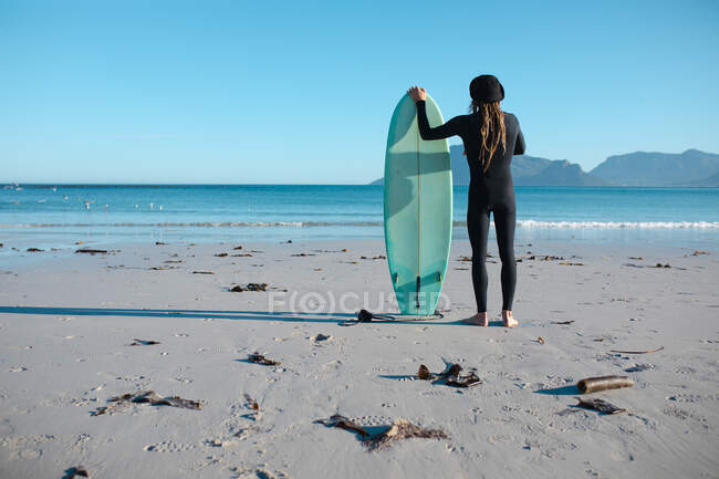 Вид сзади на человека в гидрокостюме, стоящего с доской для серфинга, смотрящего на чистое небо с копировальным пространством с пляжа. хобби и водные виды спорта. — стоковое фото