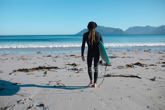 Vista posteriore dell'uomo in muta che cammina con la tavola da surf in spiaggia verso lo spazio copia sul cielo blu. hobby e sport acquatici. — Foto stock