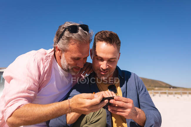 Feliz pareja gay caucásica abrazando y utilizando el teléfono inteligente por un coche en la playa. viaje por carretera de verano y vacaciones en la naturaleza. - foto de stock