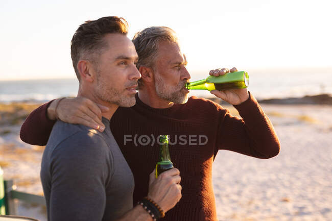 Feliz pareja gay caucásica bebiendo botellas de cerveza en un día soleado en la playa. viaje por carretera de verano y vacaciones en la naturaleza. - foto de stock
