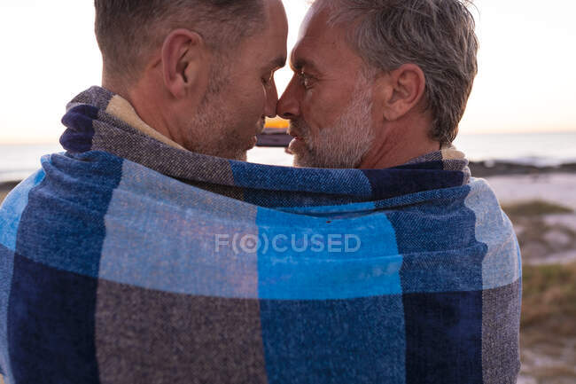 Feliz casal masculino gay caucasiano envolto em cobertor, abraçando e tirando selfie na praia ao pôr do sol. viagem de verão e férias na natureza. — Fotografia de Stock