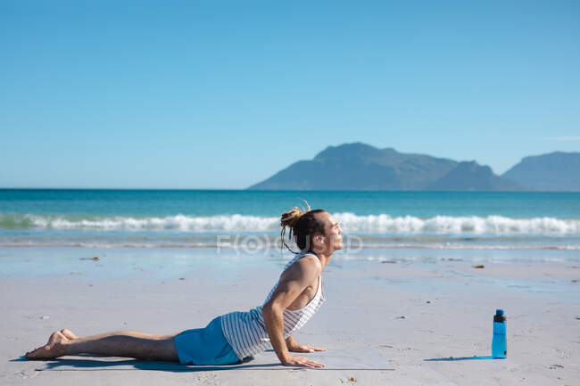 Uomo che pratica cobra posa yoga sul tappeto in spiaggia contro il cielo blu con spazio copia. fitness e stile di vita sano. — Foto stock