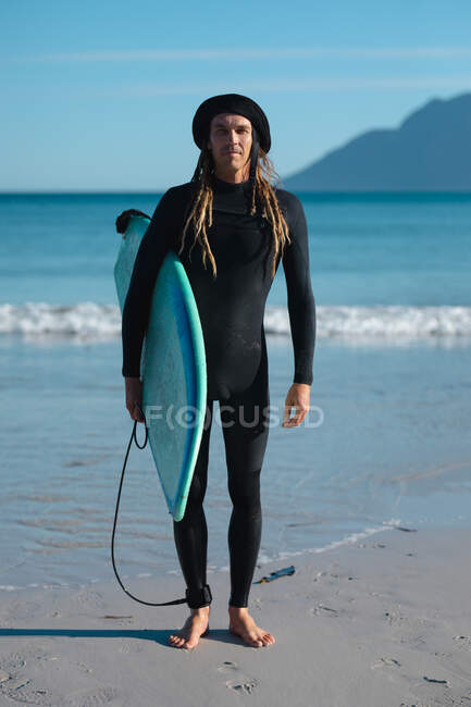 Portrait d'un surfeur masculin confiant en combinaison noire portant une planche de surf à la plage par une journée ensoleillée. hobbies et sports nautiques. — Photo de stock