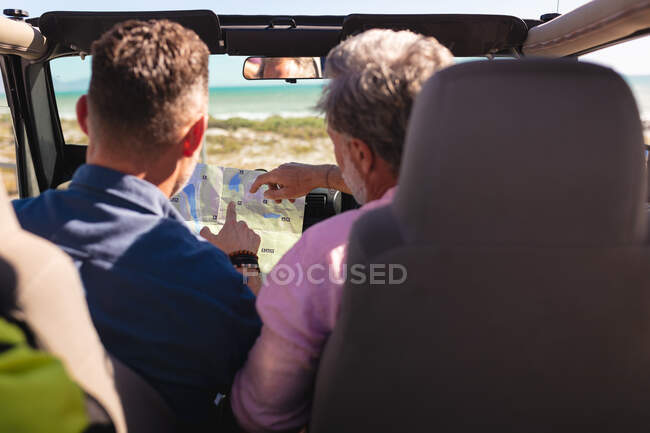 Caucasian gay male couple reading map sitting in car at seaside. summer road trip and holiday in nature. — Stock Photo