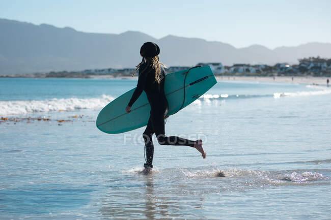 Longitud completa de hipster masculino corriendo con tabla de surf en la playa durante el fin de semana. pasatiempos y deportes acuáticos. - foto de stock