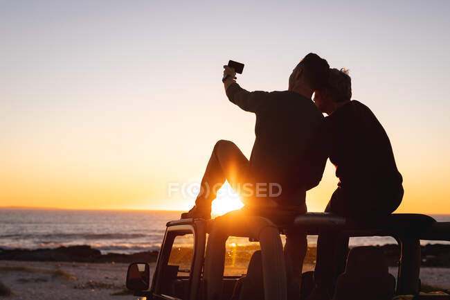 Rear view of caucasian gay male couple sitting on car roof taking selfies at sunset by the sea. summer road trip and holiday in nature. — Stock Photo