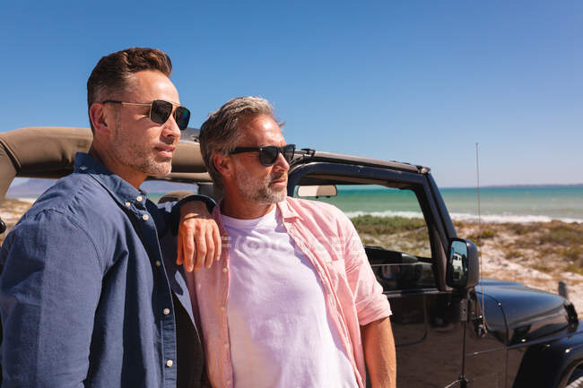 Happy caucasian gay male couple embracing by a car at seaside. summer road trip and holiday in nature. — Stock Photo