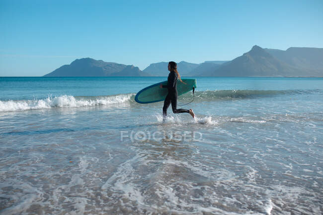 Man carrying surfboard running while splashing water on shore against blue sky with copy space. hobbies and water sport. — Stock Photo