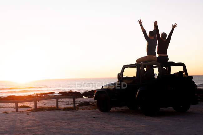 Caucasian gay male couple sitting on car roof raising arms and holding hands at sunset by the sea. summer road trip and holiday in nature. — Stock Photo
