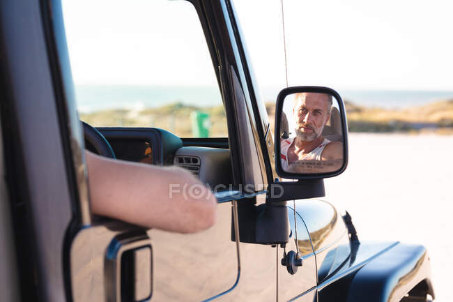 Porträt eines lächelnden kaukasischen Mannes im Auto, das sich an einem sonnigen Tag am Meer im Seitenspiegel spiegelt. Sommer Roadtrip und Urlaub in der Natur. — Stockfoto