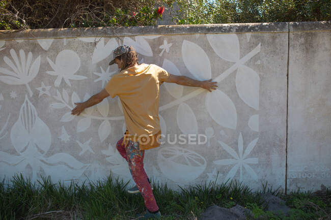 Vista trasera del artista masculino tocando pintura mural abstracta en la pared mientras camina sobre hierba. arte urbano y habilidad. - foto de stock