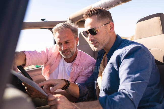Feliz pareja gay caucásica leyendo mapa sentado en el coche a la orilla del mar. viaje por carretera de verano y vacaciones en la naturaleza. - foto de stock