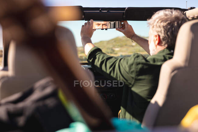 Caucasian man adjusting rearview mirror in car at seaside. summer road trip and holiday in nature. — Stock Photo