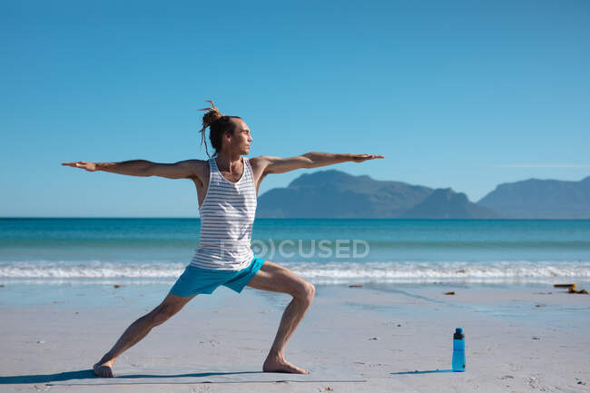 Hombre practicante de guerrero 2 pose yoga con los brazos extendidos en la playa contra el cielo con espacio de copia. fitness y estilo de vida saludable. - foto de stock