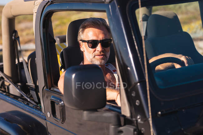 Portrait of caucasian man wearing sunglasses sitting in car on sunny day at seaside. summer road trip and holiday in nature. — Stock Photo