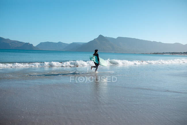 Surfista corriendo en la orilla con tabla de surf en la playa contra el cielo azul claro y espacio de copia. pasatiempos y deportes acuáticos. - foto de stock