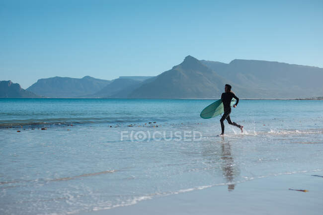 Male surfer carrying surfboard while running on shore splashing water against copy space. hobbies and water sport. — Stock Photo