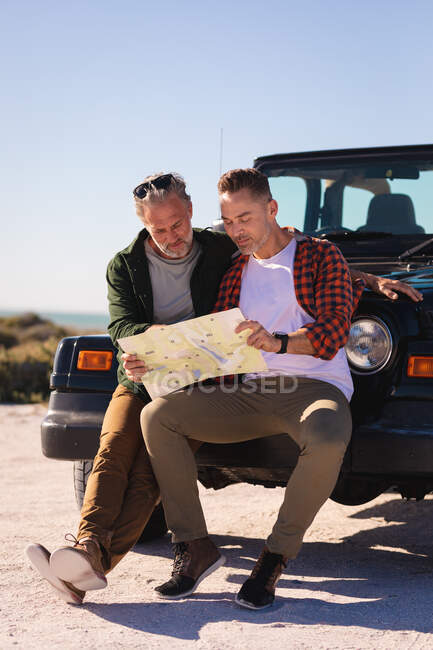 Feliz pareja gay caucásica leyendo mapa apoyado en el coche en la playa. viaje por carretera de verano y vacaciones en la naturaleza. - foto de stock