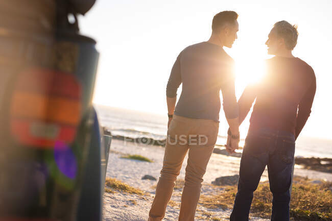 Happy caucasian gay male couple holding hands, walking away from car at seaside. summer road trip and holiday in nature. — Stock Photo