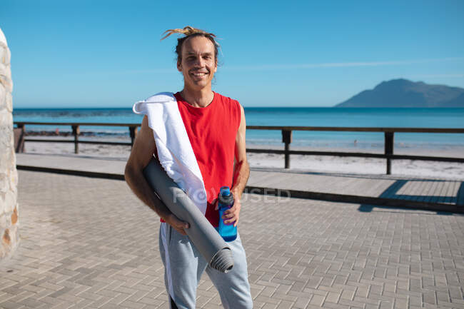 Retrato de homem sorridente segurando tapete de ioga e garrafa com toalha no ombro na praia. fitness e estilo de vida saudável. — Fotografia de Stock