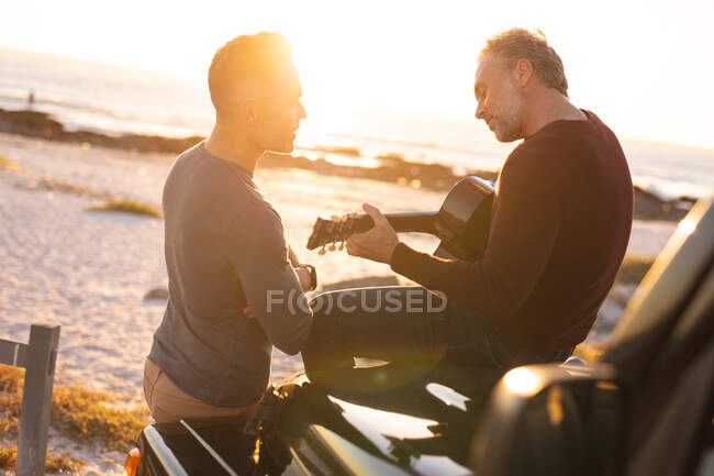 Щаслива біла пара геїв, що висить на пляжі на світанку, сидить на машині, граючи на гітарі. літня дорога поїздка і відпочинок на природі . — стокове фото