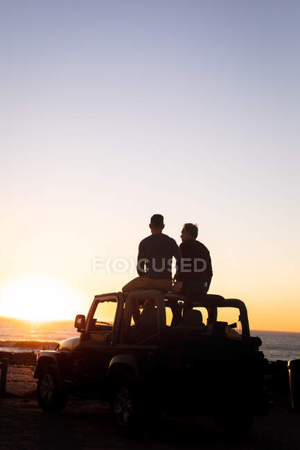Rear view of caucasian gay male couple sitting on car roof at sunset by the sea. summer road trip and holiday in nature. — Stock Photo