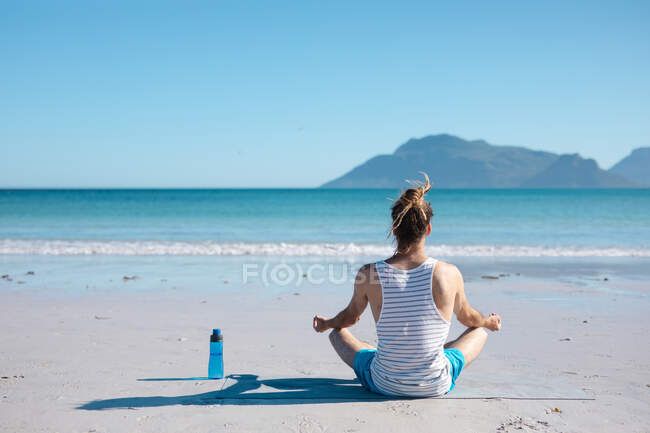 Задний вид человека, медитирующего во время занятий йогой на пляже с копировальным пространством в ясном голубом небе. фитнес и здоровый образ жизни. — стоковое фото