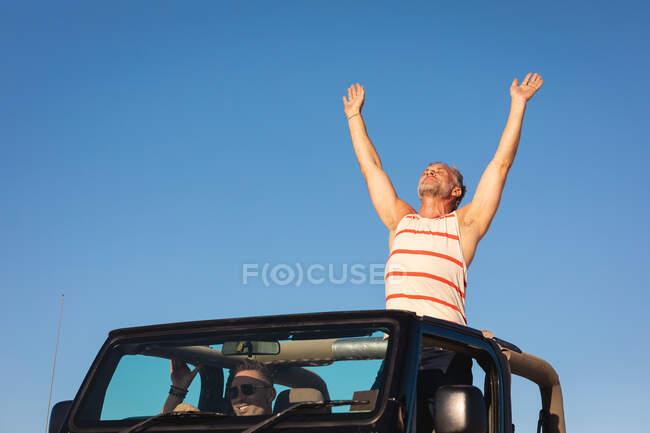 Feliz pareja gay caucásica levantando brazos sentados en coche en un día soleado en la playa. viaje por carretera de verano y vacaciones en la naturaleza. - foto de stock