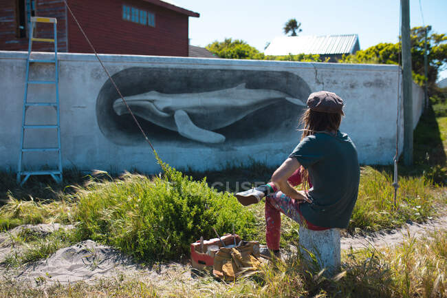 Artista masculino sentado enquanto admira seu belo mural de baleia na parede durante o dia ensolarado. arte de rua e habilidade. — Fotografia de Stock