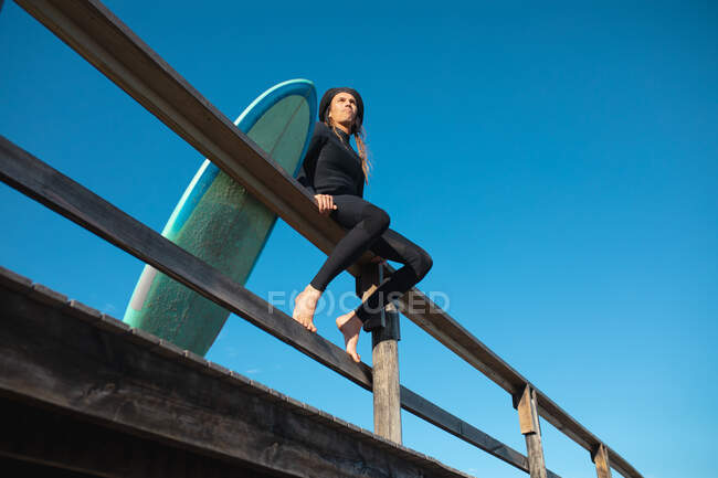 Low angle view of man sitting with surfboard on railing against clear blue sky with copy space. hobbies and water sport. — Stock Photo