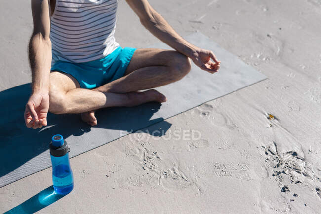 Low section of man meditating while practicing yoga by water bottle on sand at beach. fitness and healthy lifestyle. — Stock Photo