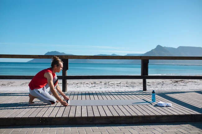 Mann kniet beim Auslegen von Yogamatte für das Training auf Dielenbrett am Strand gegen den Himmel mit Kopierraum. Fitness und gesunder Lebensstil. — Stockfoto