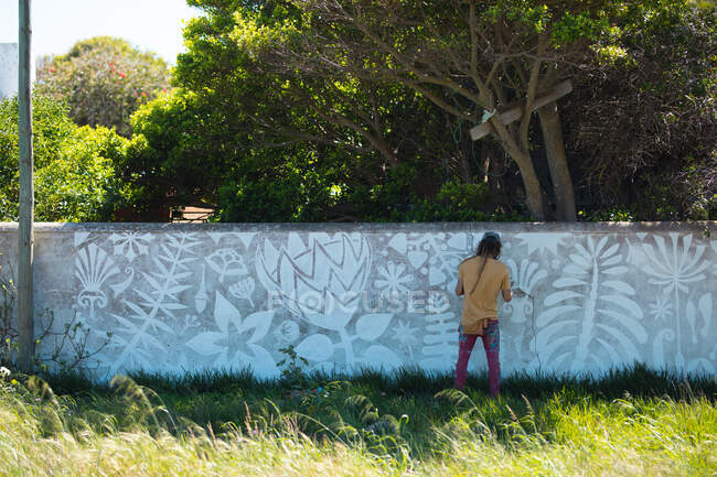 Vista trasera de cuerpo entero del artista masculino sosteniendo la paleta mientras pinta un mural abstracto en la pared. arte urbano y habilidad. - foto de stock