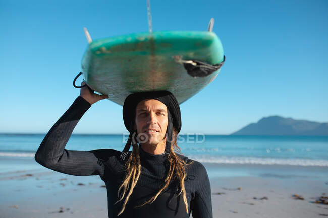 Retrato del hombre hipster llevando tabla de surf en la cabeza en la playa contra el cielo azul. pasatiempos y deportes acuáticos. - foto de stock