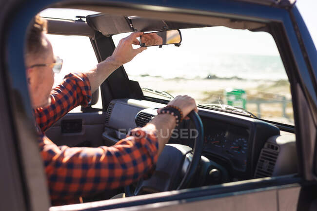 Hombre caucásico ajustando espejo retrovisor en coche a la orilla del mar. viaje por carretera de verano y vacaciones en la naturaleza. - foto de stock