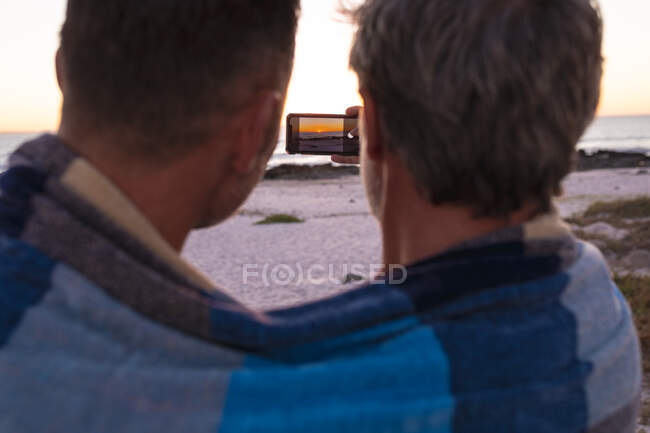 Heureux couple gay caucasien enveloppé dans une couverture, embrassant et prenant selfie sur la plage au coucher du soleil. road trip d'été et vacances dans la nature. — Photo de stock