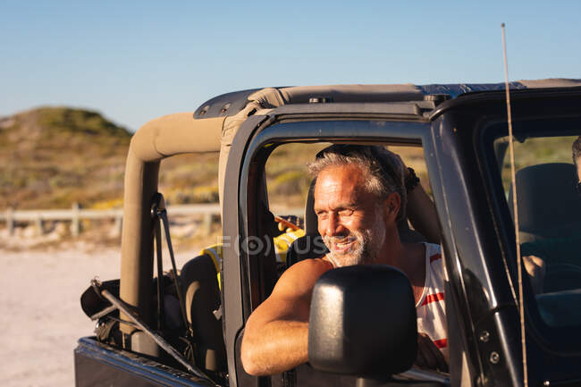 Happy caucasian man sitting in car smiling on sunny day at seaside. summer road trip and holiday in nature. — Stock Photo