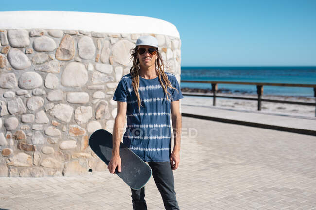 Portrait of male hipster holding skateboard against wall at promenade on sunny day. lifestyle and sport. — Stock Photo