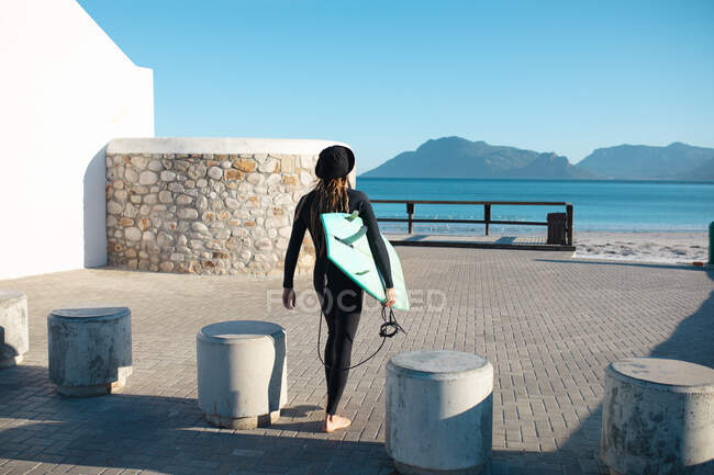 Vista trasera del surfista hombre llevando tabla de surf caminando en medio de pilonas de hormigón en el día soleado. pasatiempos y deportes acuáticos. - foto de stock