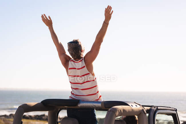 Rear view of caucasian man raising arms sitting on car on sunny day at the seaside. summer road trip and holiday in nature. — Stock Photo