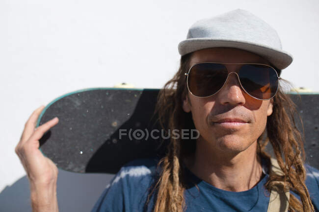 Porträt eines modischen Hipster-Mannes mit Sonnenbrille und Schirmmütze und Skateboard hinter dem Kopf. Lifestyle, Mode und Sport. — Stockfoto