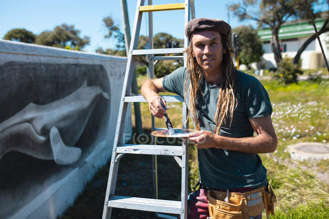 Porträt eines männlichen Künstlers, der Pinsel und Palette an die Wand hält und sich auf eine Leiter lehnt. Street Art und Geschicklichkeit. — Stockfoto