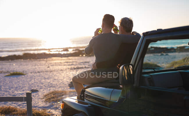 Счастливая кавказская гей-пара пьет пиво, сидя на машине на закате у моря. летняя поездка и отдых на природе. — стоковое фото