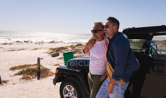 Happy caucasian gay male couple embracing and enjoying the view by car at seaside. summer road trip and holiday in nature. — Stock Photo
