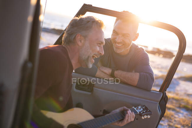 Glückliches kaukasisches schwules Männerpaar, das Gitarre spielt und lacht und mit dem Auto am Meer abhängt. Sommer Roadtrip und Urlaub in der Natur. — Stockfoto