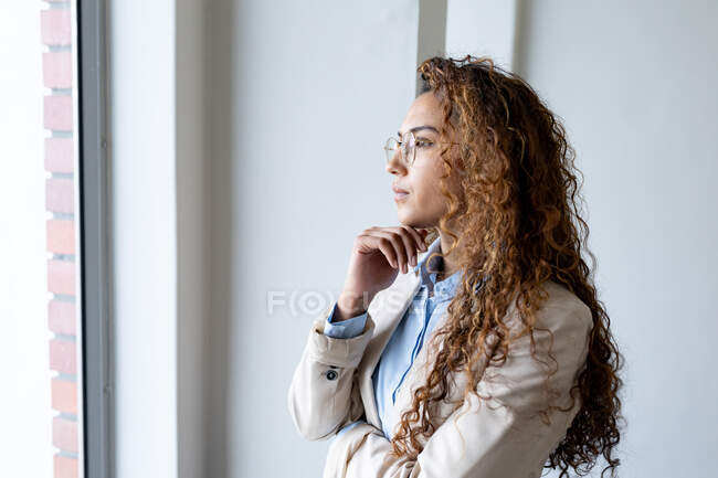Pensativa mujer de negocios birracial mirando a través de la ventana mientras está de pie con la mano en la barbilla en la oficina. negocios y oficina de trabajo. - foto de stock
