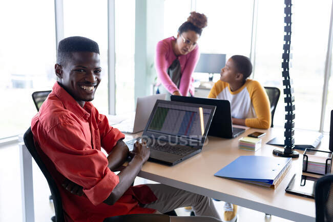 Retrato del sonriente hombre de negocios afroamericano con portátil en el escritorio en la oficina creativa. negocio creativo, tecnología inalámbrica y lugar de trabajo de oficina. - foto de stock