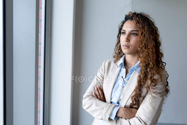 Biracial businesswoman looking away while standing with arms crossed in office. business and office workplace. — Stock Photo
