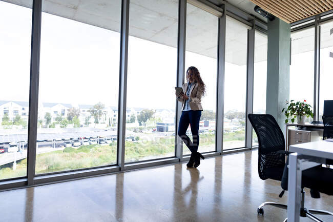 Kreative Geschäftsfrau mit digitalem Tablet, während sie sich im Büro ans Fenster lehnt. Business, Büroarbeitsplätze und drahtlose Technologie. — Stockfoto