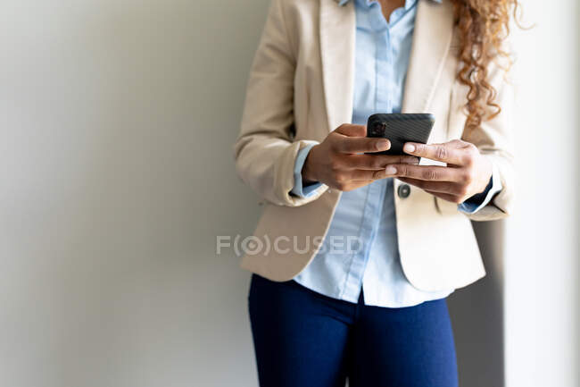 Midsection de la mujer de negocios que usa casuales inteligentes usando el teléfono inteligente contra la pared en la oficina. Negocios, trabajo de oficina y tecnología inalámbrica. - foto de stock